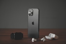 A sleek, modern smartphone with a dark metallic finish stands upright on a wooden surface. To its left lies a black smartwatch, while wireless earbuds and their open charging case are positioned to its right. The scene is set against a muted dark background, highlighting the elegance of the gadgets.