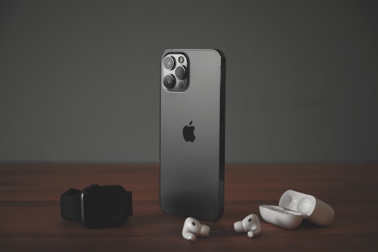 A sleek, modern smartphone with a dark metallic finish stands upright on a wooden surface. To its left lies a black smartwatch, while wireless earbuds and their open charging case are positioned to its right. The scene is set against a muted dark background, highlighting the elegance of the gadgets.