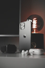 A sleek smartphone with a metallic finish stands upright on a table alongside a modern smartwatch, wireless earbuds, and a glowing antique-style bulb. The scene is set against a dark background, emphasizing the shiny surfaces and elegant technology.