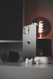A sleek smartphone with a metallic finish stands upright on a table alongside a modern smartwatch, wireless earbuds, and a glowing antique-style bulb. The scene is set against a dark background, emphasizing the shiny surfaces and elegant technology.