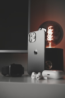 A sleek smartphone with a metallic finish stands upright on a table alongside a modern smartwatch, wireless earbuds, and a glowing antique-style bulb. The scene is set against a dark background, emphasizing the shiny surfaces and elegant technology.