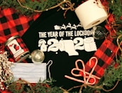 A black t-shirt with 'The Year of the Lockdown 2020' text in white, featuring images of toilet paper rolls and a Santa hat. Surrounding items include a roll of toilet paper, a face mask, candy canes, a red and black checkered Michigan-themed candle, two transparent Christmas ornaments, pine branches, and a rustic rope.