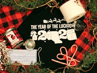 A black t-shirt with 'The Year of the Lockdown 2020' text in white, featuring images of toilet paper rolls and a Santa hat. Surrounding items include a roll of toilet paper, a face mask, candy canes, a red and black checkered Michigan-themed candle, two transparent Christmas ornaments, pine branches, and a rustic rope.