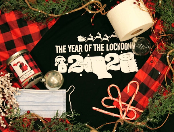 A black t-shirt with 'The Year of the Lockdown 2020' text in white, featuring images of toilet paper rolls and a Santa hat. Surrounding items include a roll of toilet paper, a face mask, candy canes, a red and black checkered Michigan-themed candle, two transparent Christmas ornaments, pine branches, and a rustic rope.