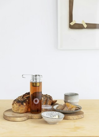 A neatly arranged breakfast setting with a loaf of sliced bread on a wooden board. A glass bottle filled with amber-colored tea is placed centrally among the items. Various ceramic dishes contain butter, dried fruits, and a cup, complementing the rustic theme. A minimalistic white background with a framed art piece adds elegance to the composition.
