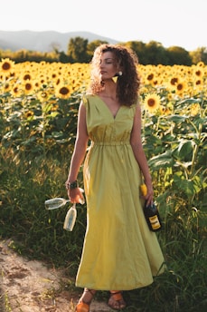 A woman stands in a lush field of sunflowers under a clear sky. She wears a long, light green dress and holds two empty wine glasses and a bottle of wine. Her curly hair is illuminated by the sunlight, and a mountain range is visible in the background.