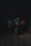 A person with a backpack walking alone through a dimly lit alley or passage at night. The environment is shadowy and subdued, creating a sense of isolation.