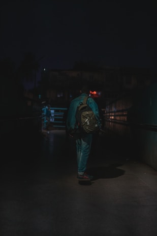 A person with a backpack walking alone through a dimly lit alley or passage at night. The environment is shadowy and subdued, creating a sense of isolation.