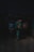 A person with a backpack walking alone through a dimly lit alley or passage at night. The environment is shadowy and subdued, creating a sense of isolation.