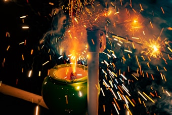Bright sparks and flames erupt from a small firework placed inside a green ceramic pot, surrounded by colorful trails of light and smoke creating a dynamic and festive scene.