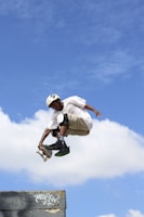 A skateboarder is performing a high-flying trick in the air against a backdrop of a clear blue sky with some white clouds. The skateboarder is wearing a helmet, knee pads, and casual clothing, indicating an emphasis on safety while engaging in the sport.