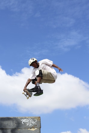 A skateboarder is performing a high-flying trick in the air against a backdrop of a clear blue sky with some white clouds. The skateboarder is wearing a helmet, knee pads, and casual clothing, indicating an emphasis on safety while engaging in the sport.