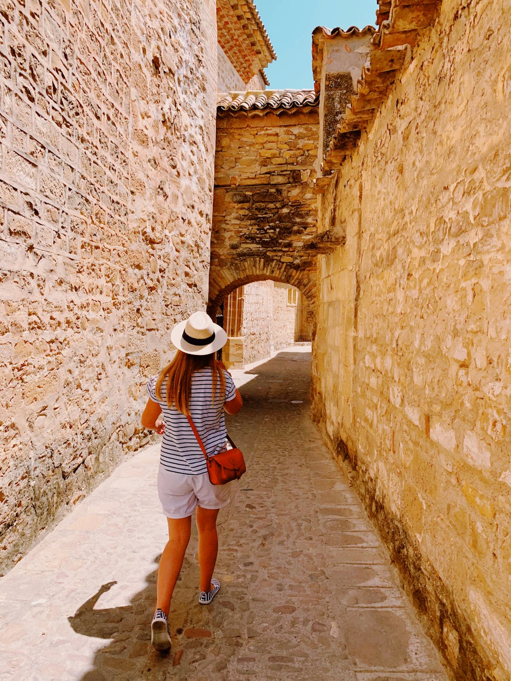 woman in white and black striped shirt and blue denim shorts walking on brown concrete pathway