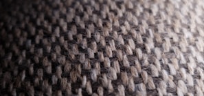 A close-up view of a textured woven fabric featuring a pattern of interlaced threads in shades of brown and cream. The fibers appear slightly fuzzy, giving the surface a soft and tactile appearance.