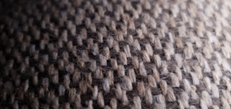 A close-up view of a textured woven fabric featuring a pattern of interlaced threads in shades of brown and cream. The fibers appear slightly fuzzy, giving the surface a soft and tactile appearance.