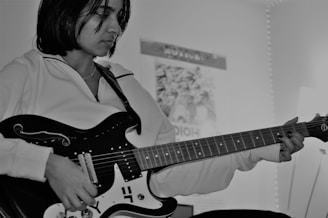 A person is playing an electric guitar, focusing intently on the instrument. The environment suggests a personal space with a poster on the wall and soft lighting in the background. The image is in black and white, giving it a classic and introspective feel.