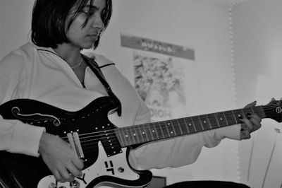 A person is playing an electric guitar, focusing intently on the instrument. The environment suggests a personal space with a poster on the wall and soft lighting in the background. The image is in black and white, giving it a classic and introspective feel.