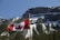 A red and white flag with a maple leaf flutters against the backdrop of a rugged mountain range. The mountains are covered with patches of evergreen trees and snow, under a clear blue sky.