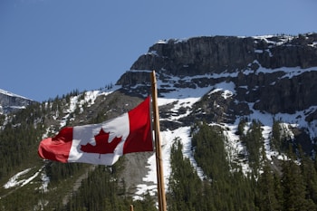 A red and white flag with a maple leaf flutters against the backdrop of a rugged mountain range. The mountains are covered with patches of evergreen trees and snow, under a clear blue sky.