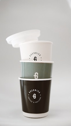 Three stacked coffee cups with minimalist designs, each labeled with 'Guernica Coffee & Gallery'. The cups are in varying shades of white, gray, and black, arranged vertically with a subtle gradient effect.