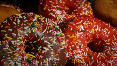 A close-up view of colorful donuts topped with a generous amount of multicolored sprinkles. The donuts have a glossy appearance and vibrant icing, including shades of chocolate brown and bright red.