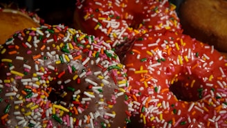 A close-up view of colorful donuts topped with a generous amount of multicolored sprinkles. The donuts have a glossy appearance and vibrant icing, including shades of chocolate brown and bright red.