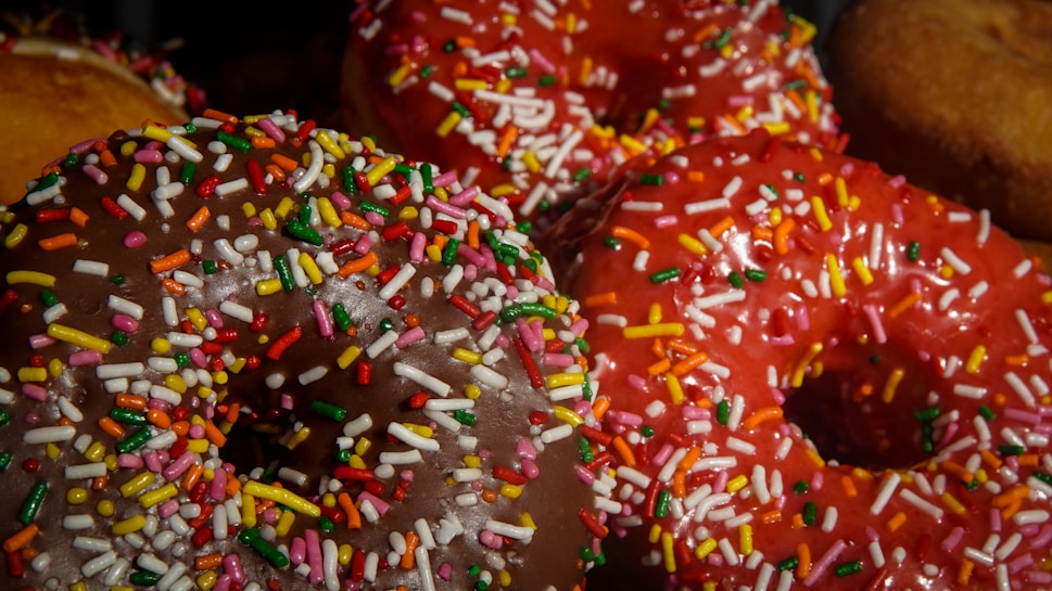 A close-up view of colorful donuts topped with a generous amount of multicolored sprinkles. The donuts have a glossy appearance and vibrant icing, including shades of chocolate brown and bright red.