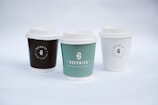 Three coffee cups with lids are arranged side by side. Each cup has a logo and the name 'Guernica Coffee & Patisserie' printed on it. The cup on the left is dark brown, the middle one is green, and the one on the right is white. The background is a soft white, creating a minimalist and clean aesthetic.