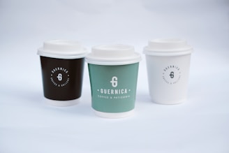 Three coffee cups with lids are arranged side by side. Each cup has a logo and the name 'Guernica Coffee & Patisserie' printed on it. The cup on the left is dark brown, the middle one is green, and the one on the right is white. The background is a soft white, creating a minimalist and clean aesthetic.