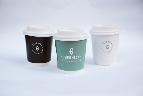 Three coffee cups with lids are arranged side by side. Each cup has a logo and the name 'Guernica Coffee & Patisserie' printed on it. The cup on the left is dark brown, the middle one is green, and the one on the right is white. The background is a soft white, creating a minimalist and clean aesthetic.