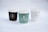 Three coffee cups with lids are arranged side by side. Each cup has a logo and the name 'Guernica Coffee & Patisserie' printed on it. The cup on the left is dark brown, the middle one is green, and the one on the right is white. The background is a soft white, creating a minimalist and clean aesthetic.