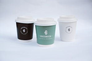 Three coffee cups with lids are arranged side by side. Each cup has a logo and the name 'Guernica Coffee & Patisserie' printed on it. The cup on the left is dark brown, the middle one is green, and the one on the right is white. The background is a soft white, creating a minimalist and clean aesthetic.