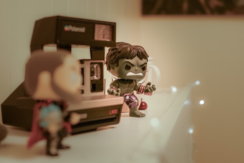 A shelf displaying a vintage Polaroid camera alongside two superhero action figures, one prominently in focus with a green muscular appearance and a fierce expression. The background has warm lighting and soft bokeh effects.