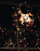 A vividly decorated arrangement with various ornaments, including a prominent illuminated snowflake in the center. The decorations include red and gold baubles, surrounded by twinkling fairy lights that cast a warm glow. The background is dark, enhancing the brightness and colors of the decorations.