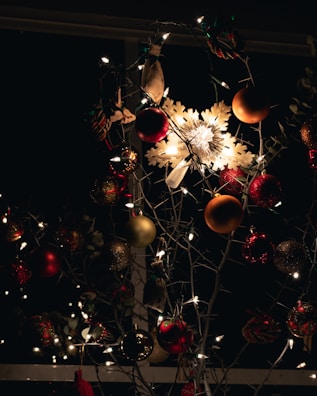A vividly decorated arrangement with various ornaments, including a prominent illuminated snowflake in the center. The decorations include red and gold baubles, surrounded by twinkling fairy lights that cast a warm glow. The background is dark, enhancing the brightness and colors of the decorations.