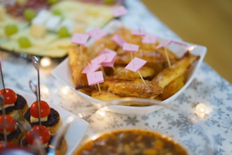 A close-up view of a table with assorted appetizers, including a bowl of golden pastries topped with pink flags, skewered finger foods with tomatoes, cheese, and olives, and a bowl of stew-like dish. The tablecloth has a subtle snowflake pattern and string lights add a festive touch.