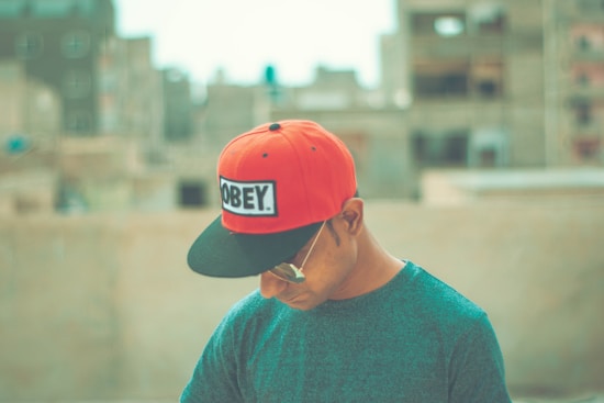 A person wearing a red and black cap with the word 'OBEY' on it is looking downwards. The background is blurred with buildings, suggesting an urban setting. The person is also wearing sunglasses and a dark-colored shirt.