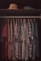 A collection of patterned shirts hanging neatly on hangers in a closet. Each shirt features a distinct and colorful design, ranging from floral prints to geometric patterns. Above the shirts, two hats are placed on a shelf, one straw hat and one that appears to be a flat cap.