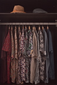 A collection of patterned shirts hanging neatly on hangers in a closet. Each shirt features a distinct and colorful design, ranging from floral prints to geometric patterns. Above the shirts, two hats are placed on a shelf, one straw hat and one that appears to be a flat cap.