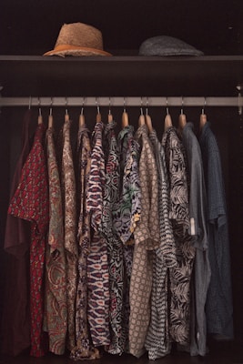 A collection of patterned shirts hanging neatly on hangers in a closet. Each shirt features a distinct and colorful design, ranging from floral prints to geometric patterns. Above the shirts, two hats are placed on a shelf, one straw hat and one that appears to be a flat cap.