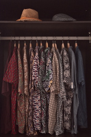 A collection of patterned shirts hanging neatly on hangers in a closet. Each shirt features a distinct and colorful design, ranging from floral prints to geometric patterns. Above the shirts, two hats are placed on a shelf, one straw hat and one that appears to be a flat cap.
