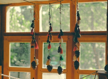 Colorful tassels and pinecones hang decoratively in front of a wooden window, with sunlight filtering through the glass and casting a warm glow.