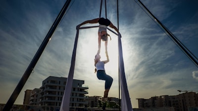 Two people are performing an aerial silk routine against a backdrop of an urban landscape with several buildings. The sky is clear with a few clouds and the sun is setting or rising behind them, casting a dramatic light and shadow effect on the scene.