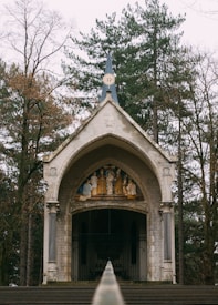 A small stone chapel situated in a wooded area, featuring a pointed blue spire and decorative reliefs above the entrance. Tall trees surround the structure, contributing to a serene and secluded ambiance.