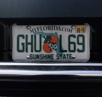 A Florida license plate with the numbers and letters 'GHUL69'. It features the outline of the state and an orange graphic with blossoms. The text 'SUNSHINE STATE' is displayed at the bottom with the website 'MYFLORIDA.COM' at the top. There is a yellow sticker in the upper right corner indicating an expiration date of 06-19.