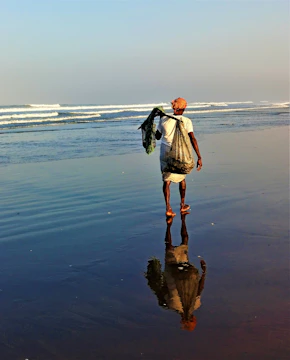 An elderly man standing barefoot on a rocky shore, gazing at the horizon under a soft sunset.