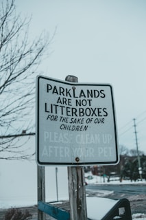 A weathered sign stands prominently, warning visitors that parklands are not to be treated as litterboxes for the sake of children. The bottom of the sign reminds readers to clean up after their pets. It is mounted on a wooden post and set against a background of bare trees and a snowy landscape, possibly in an urban setting.