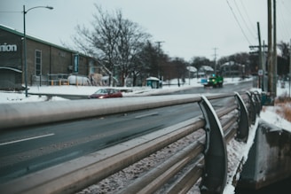 A snowy urban scene with a road flanked by metal guardrails and a building in the background. Two vehicles, a red car and a green truck, are seen on the road. The trees appear leafless, indicating winter, and the cloudy sky adds to the chilly atmosphere.