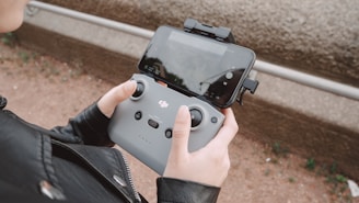 A person is holding a DJI remote controller attached to a smartphone. The controller is white with black buttons and joysticks, and the smartphone displays a screen with various controls visible. The person is wearing a black jacket and is standing outside on a dirt path with some greenery in the background.