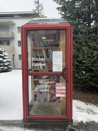 A small, red-framed library kiosk situated outdoors. Its shelves are filled with a variety of books visible through the glass door, which displays German text. The ground is snowy, and there are evergreen trees nearby, along with a modern building in the background.